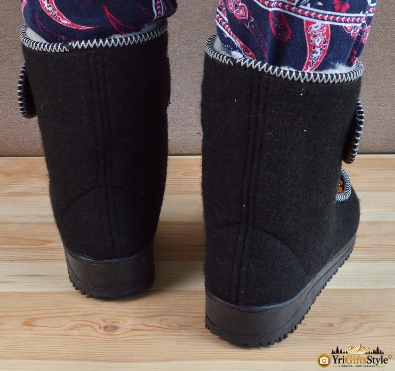 May include: A pair of black felt boots featuring a white zigzag stitch along the top edge, a black rubber sole, and a side closure strap. The interior is lined with white fleece, and the boots are worn with dark blue pants with a red paisley pattern.