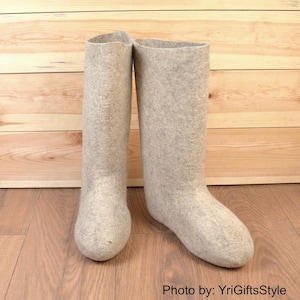 Könnte beinhalten: Ein Paar hellgraue Filzstiefel. Die Stiefel sind hoch, reichen bis zur Mitte der Wade und haben ein schlichtes, abgerundetes Zehendesign. Die Stiefel sind aus einem dicken, weichen Material gefertigt und stehen auf einer Holzoberfläche.