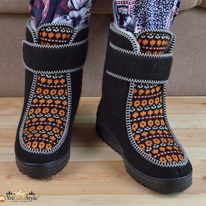 May include: A pair of black mid-calf boots featuring a textured orange, white, and black geometric knit pattern on the front panel. The boots have a black hook-and-loop strap closure at the top and a thick black rubber sole with white stitching.