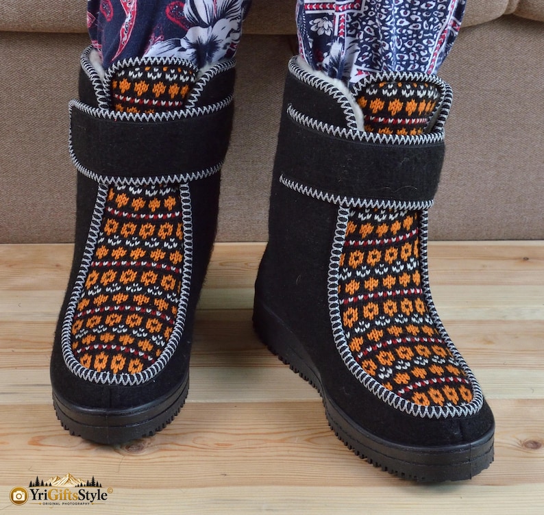 May include: A pair of black mid-calf boots featuring a textured orange, white, and black geometric knit pattern on the front panel. The boots have a black hook-and-loop strap closure at the top and a thick black rubber sole with white stitching.
