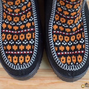 May include: A pair of black boots featuring a central panel with a vibrant orange, white, and black geometric knit pattern. The panel is outlined with a thick, light-colored decorative whipstitch detail, contrasting against the solid black material.