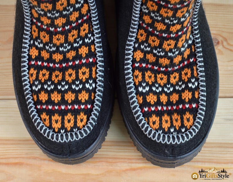 May include: A pair of black boots featuring a central panel with a vibrant orange, white, and black geometric knit pattern. The panel is outlined with a thick, light-colored decorative whipstitch detail, contrasting against the solid black material.