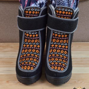 May include: A pair of black felt boots featuring a central panel with a multi-colored orange, white, and black geometric knit pattern. The boots have a thick black rubber sole, a white stitched trim, and a wide black hook-and-loop closure strap.