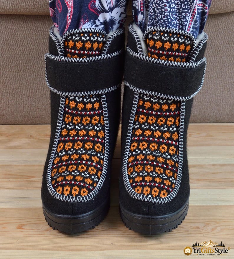 May include: A pair of black felt boots featuring a central panel with a multi-colored orange, white, and black geometric knit pattern. The boots have a thick black rubber sole, a white stitched trim, and a wide black hook-and-loop closure strap.
