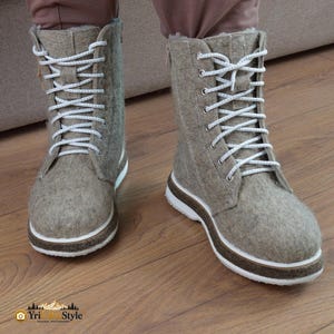 Könnte beinhalten: Ein Paar wadenhohe Stiefel aus hellbrauner Filzwolle. Die Stiefel verfügen über eine Schnürung mit weißen Schnürsenkeln, silberfarbene Metallösen und eine dicke, mehrschichtige Sohle in Weiß und Braun. Mit seitlichem Reißverschluss.