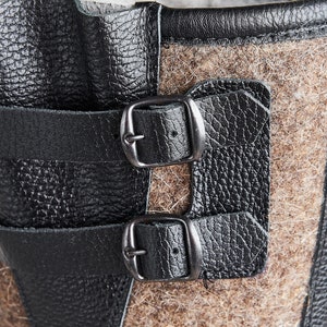 Puede incluir: Primer plano de un par de correas de cuero negro con hebillas plateadas, unidas a una bota de fieltro marr&oacute;n.