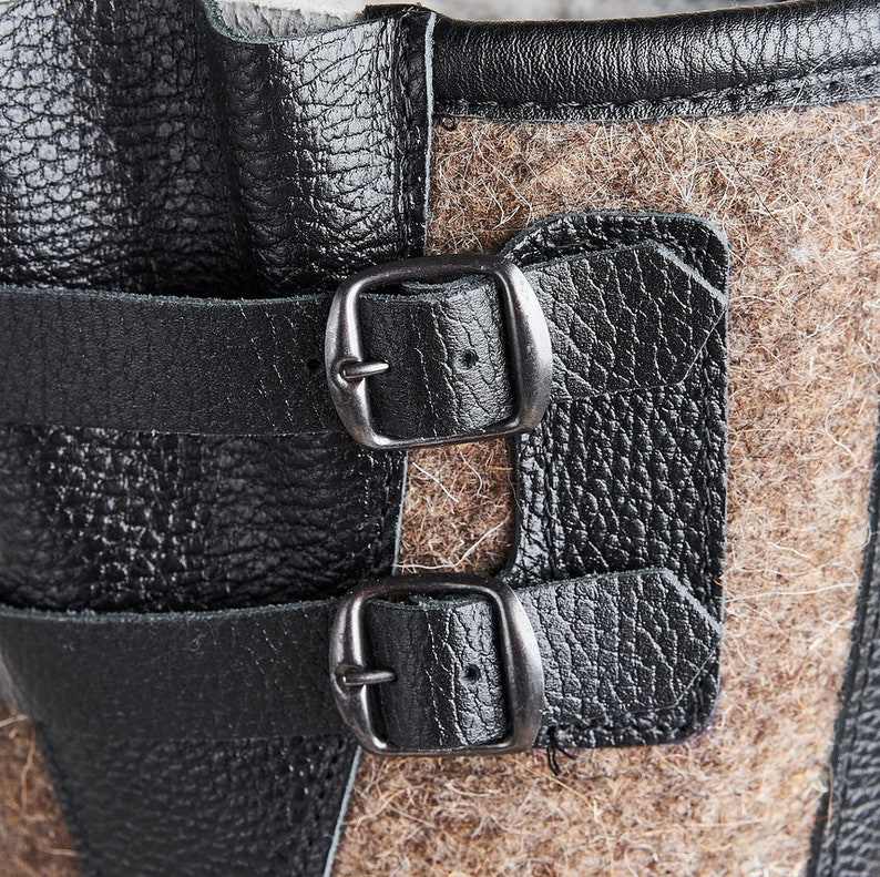 Puede incluir: Primer plano de un par de correas de cuero negro con hebillas plateadas, unidas a una bota de fieltro marr&oacute;n.