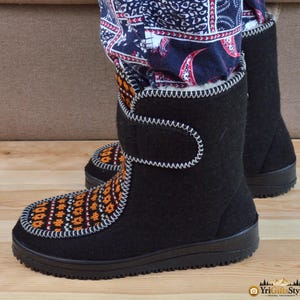 May include: Black mid-calf boots featuring a white whipstitch trim along the seams and a hook-and-loop closure strap. The front panel displays a repeating orange, black, and white geometric pattern. The boots have a black rubber sole.
