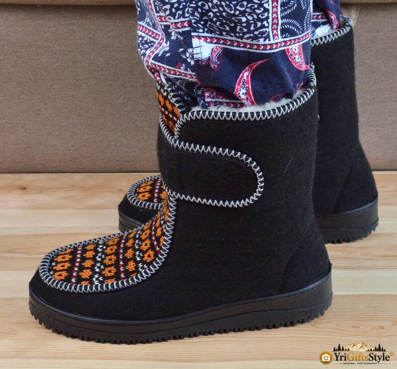 May include: Black mid-calf boots featuring a white whipstitch trim along the seams and a hook-and-loop closure strap. The front panel displays a repeating orange, black, and white geometric pattern. The boots have a black rubber sole.
