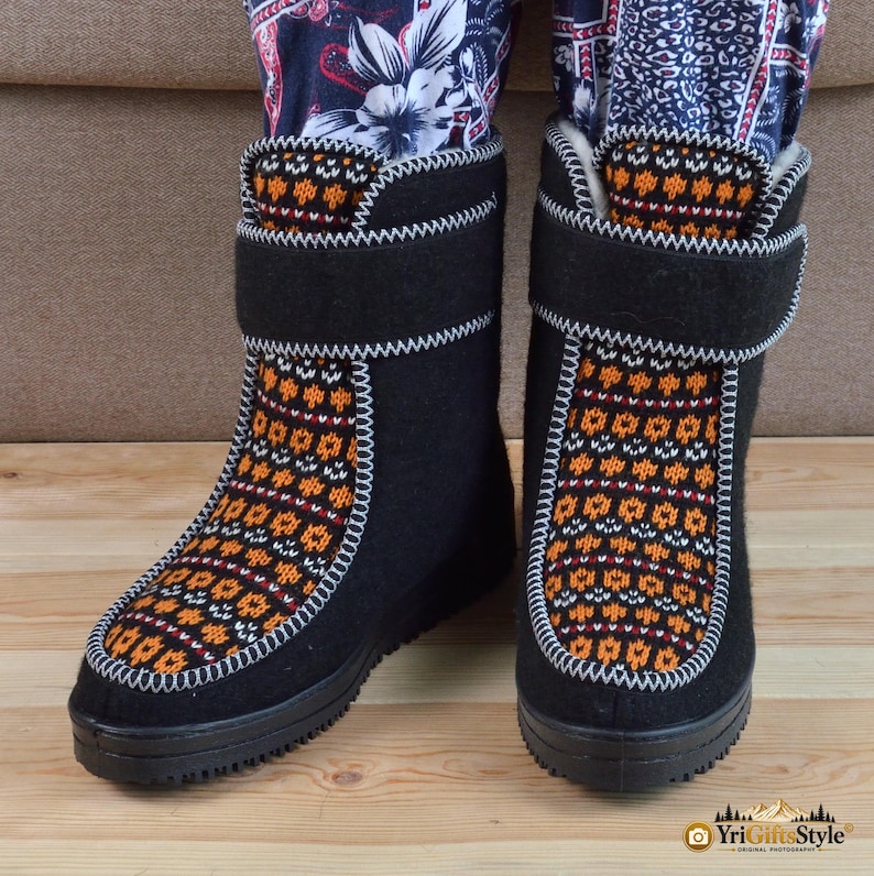 May include: A pair of black felt boots featuring a vibrant orange and white geometric knit pattern on the front panel and collar. The boots have a thick black sole, a wide hook-and-loop closure strap across the ankle, and white decorative stitching.
