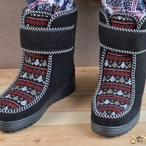 May include: A pair of black mid-calf boots featuring a central panel with a red, white, and grey geometric fair isle pattern. The boots have a thick black sole, a hook-and-loop strap closure across the ankle, and visible white whipstitch detailing.