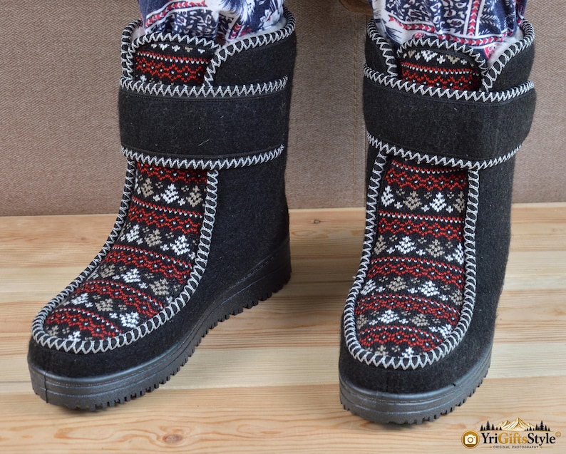 May include: A pair of black mid-calf boots featuring a central panel with a red, white, and grey geometric fair isle pattern. The boots have a thick black sole, a hook-and-loop strap closure across the ankle, and visible white whipstitch detailing.