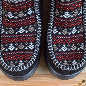 May include: A pair of black boots featuring a central panel with a knit pattern of white and tan pine trees and red geometric lines. The boots have a thick white whipstitch detail along the seam and a solid black outer border and sole.