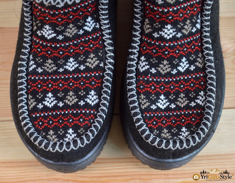May include: A pair of black boots featuring a central panel with a knit pattern of white and tan pine trees and red geometric lines. The boots have a thick white whipstitch detail along the seam and a solid black outer border and sole.