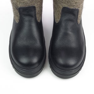 Puede incluir: Un par de botas con secciones inferiores de cuero negro texturizado y ca&ntilde;as superiores de lana afieltrada gris. Las botas tienen punteras redondeadas y suelas negras gruesas, mostrando una combinaci&oacute;n de cuero y lana.