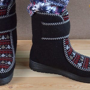 May include: A pair of black felt boots featuring a decorative panel with a red, white, and grey geometric pattern on the front. The boots have a high-top design with a hook-and-loop closure strap at the ankle and a thick black rubber sole.
