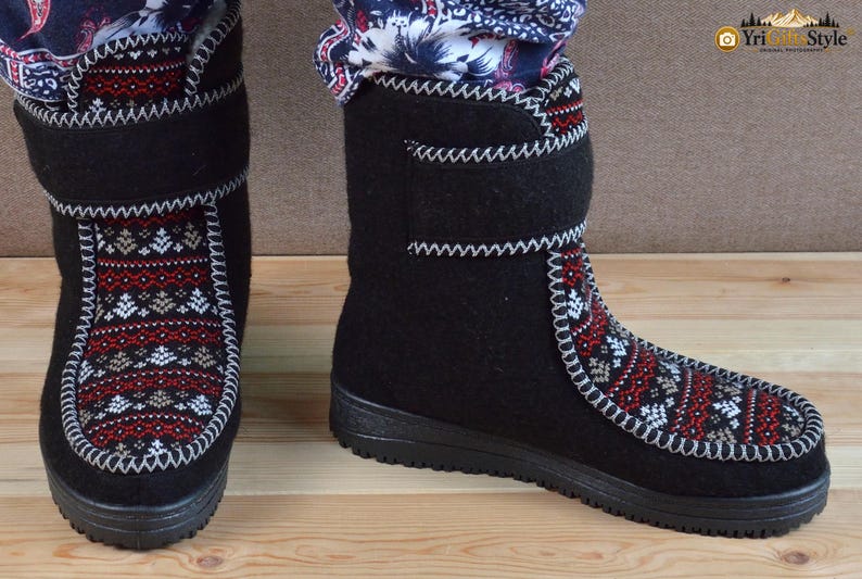 May include: A pair of black felt boots featuring a decorative panel with a red, white, and grey geometric pattern on the front. The boots have a high-top design with a hook-and-loop closure strap at the ankle and a thick black rubber sole.