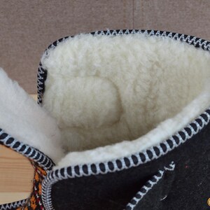 May include: A black felt boot with a thick, cream-colored wool lining. The boot features a high-top design with a wide opening, showcasing the soft interior texture. The edges are finished with a decorative black and white stitched trim.