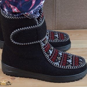 May include: Black felt boots featuring a mid-calf height, a hook-and-loop closure strap, and a black rubber sole. The front panel displays a detailed red, white, and black geometric pattern with a decorative white whipstitch trim along the edges.
