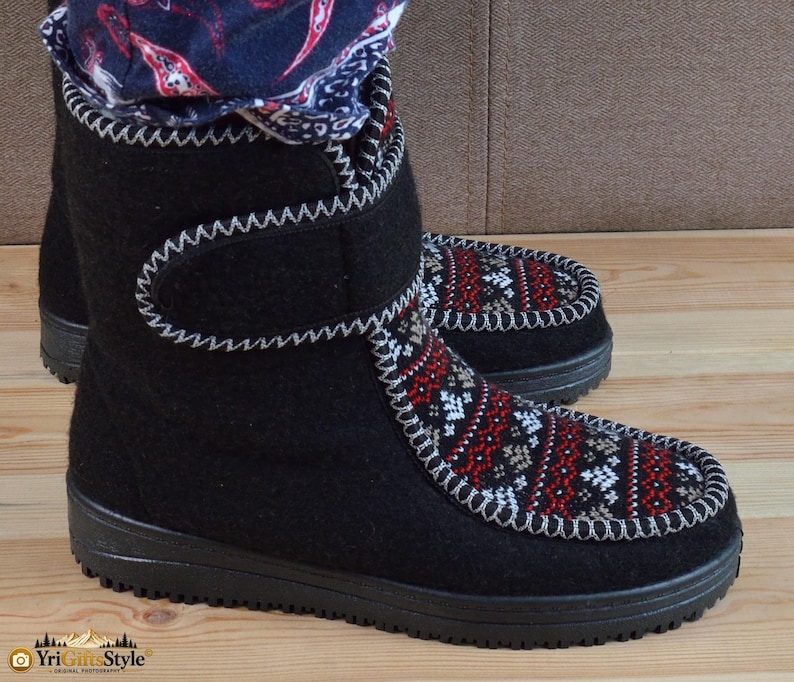 May include: Black felt boots featuring a mid-calf height, a hook-and-loop closure strap, and a black rubber sole. The front panel displays a detailed red, white, and black geometric pattern with a decorative white whipstitch trim along the edges.