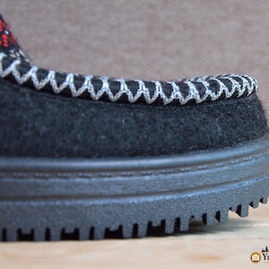 May include: A black felt shoe with a thick, black, ridged rubber sole. The upper edge features a decorative white zigzag stitch pattern, with a small section of red and white geometric embroidery visible near the top of the shoe's opening.
