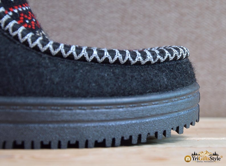 May include: A black felt shoe with a thick, black, ridged rubber sole. The upper edge features a decorative white zigzag stitch pattern, with a small section of red and white geometric embroidery visible near the top of the shoe's opening.