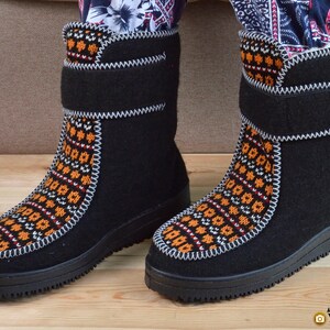 May include: A pair of black mid-calf boots featuring a central panel with an orange, white, and black geometric pattern. The boots have a thick black rubber sole, a hook-and-loop closure strap at the top, and visible white stitching along the seams.