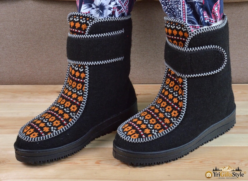 May include: A pair of black mid-calf boots featuring a central panel with an orange, white, and black geometric pattern. The boots have a thick black rubber sole, a hook-and-loop closure strap at the top, and visible white stitching along the seams.