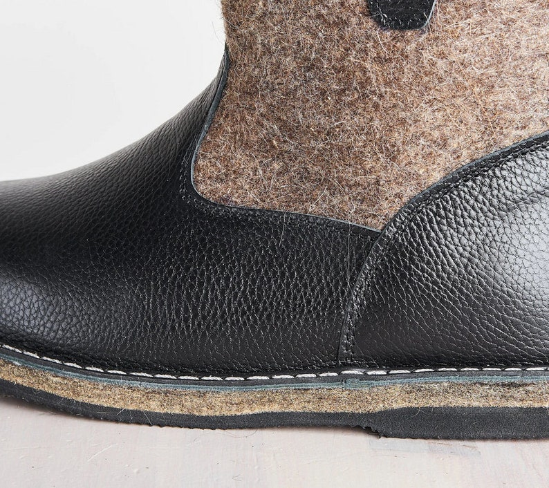 Puede incluir: Primer plano de una bota de cuero negro con una parte superior de fieltro marr&oacute;n. La bota tiene una costura cosida alrededor de la suela y una suela gruesa y texturizada.