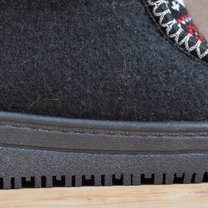 May include: A black felt boot featuring a thick, textured black rubber sole and a decorative panel on the upper section. The panel displays a geometric pattern in red, white, and black, accented by prominent white whipstitching along the edges.