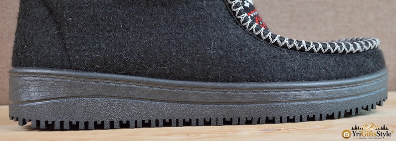 May include: A black felt boot featuring a thick, textured black rubber sole and a decorative panel on the upper section. The panel displays a geometric pattern in red, white, and black, accented by prominent white whipstitching along the edges.