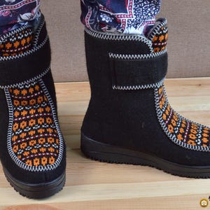 May include: A pair of black felt boots featuring a vibrant orange, white, and black geometric knit pattern on the front panel. The boots have a high-top design with a hook-and-loop closure strap at the ankle and a thick black rubber sole.
