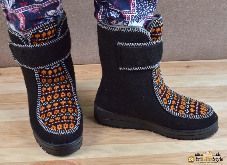 May include: A pair of black felt boots featuring a vibrant orange, white, and black geometric knit pattern on the front panel. The boots have a high-top design with a hook-and-loop closure strap at the ankle and a thick black rubber sole.