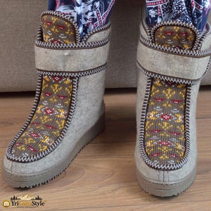 Puede incluir: Un par de botas de media caña hechas de material de fieltro gris claro. Las botas presentan un panel de punto marrón en la parte delantera con un patrón geométrico amarillo, rojo y blanco. Cada bota tiene una tira de velcro ancha y una suela marrón gruesa.
