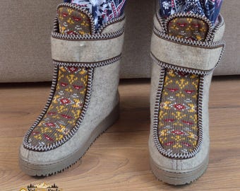 May include: A pair of mid-calf boots made of light gray felted material. The boots feature a brown knit panel on the front with a yellow, red, and white geometric pattern. Each boot has a wide hook-and-loop closure strap and a thick brown sole.