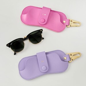 May include: Black sunglasses with gold frames next to a pink and a lilac leather glasses case. The cases have a snap closure, a gold-coloured clasp, and initials. The pink case has a heart symbol.