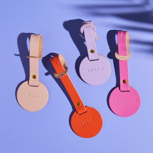 Könnte beinhalten: Vier Leder-Gepäckanhänger mit verschiedenen Namen. Die Anhänger sind in den Farben Pink, Orange, Hellrosa und Lila. Die Anhänger befinden sich auf einem hellvioletten Hintergrund.