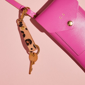 May include: A pink leather phone case with a gold clasp and the initials "C.S." embossed on the front. A gold key chain with a leopard print leather strap is attached to the case.