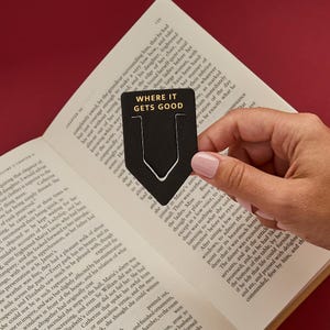 May include: A black, shield-shaped metal bookmark featuring the gold-colored text "WHERE IT GETS GOOD" printed at the top. The bookmark has a vertical cutout slot in the center and is positioned against the open pages of a printed book.