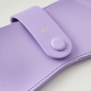 May include: A close-up of a lilac leather case with a snap closure. The case features a strap with the gold initials "AD" and a matching button. The edges are stitched with a contrasting thread.