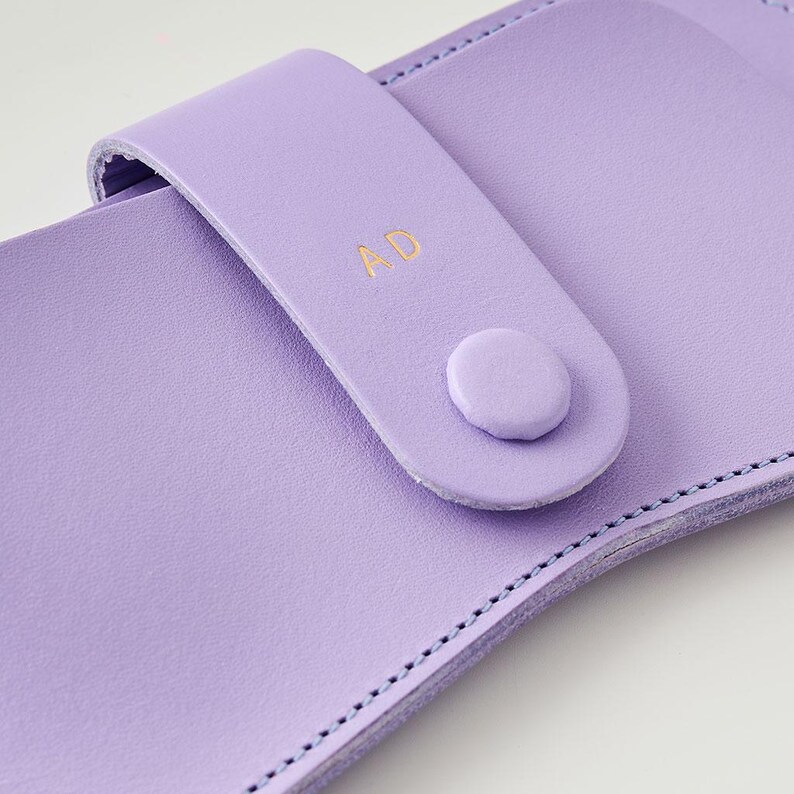 May include: A close-up of a lilac leather case with a snap closure. The case features a strap with the gold initials "AD" and a matching button. The edges are stitched with a contrasting thread.