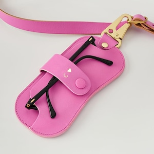May include: A vibrant pink leather glasses case with a matching strap and gold-tone clasp. The case holds black-framed glasses and has a flap with a button closure. The flap is embossed with the letter "C" and a small heart.