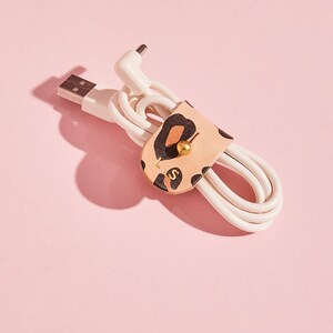 以下が含まれることがあります： レオパード柄の革製のケーブルオーガナイザーが付いた白い充電ケーブル。オーガナイザーには金色のボタンと「L.I.S.」という文字が印刷されています。