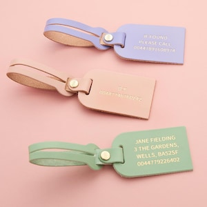 Op de afbeelding: Drie leren bagagelabels in pastelkleuren: lavendel, perzik en mintgroen. Elke tag heeft een bijpassende riem en goudkleurige hardware. De lavendelkleurige tag leest "IF FOUND PLEASE CALL 00447891608974". De perzikkleurige tag leest "D.Y. 00447746760242". De mintgroene tag leest "JANE FIELDING 3 THE GARDENS, WELLS, BA52SF 0044779226402".