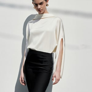 Peut inclure: Une femme porte un chemisier crème à manches ouvertes et une jupe noire longue. La jupe s'évase légèrement en bas. Elle porte des talons noirs. Le fond est un mur blanc uni.