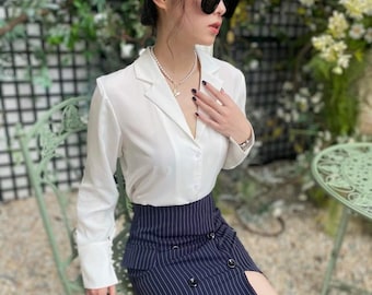 May include: A white button-down shirt with long sleeves and a navy blue pinstripe skirt with a slit on the right side. The skirt has a high waist and a button closure.