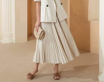 May include: A woman wearing a cream-colored linen blazer with a double-breasted closure and a matching pleated skirt. She is holding a small cream-colored clutch bag and wearing brown sandals.