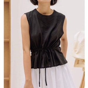 Puede incluir: Una blusa negra sin mangas con cintura de cordón y una falda blanca. La blusa tiene un escote redondo y un corte holgado.