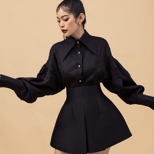 Może przedstawiać: Czarny zestaw składający się z koszuli z długim rękawem z szerokim kołnierzem i spódnicy z wysokim stanem. Koszula ma złote guziki, a spódnica ma rozkloszowaną sylwetkę. Zestaw jest na neutralnym tle.