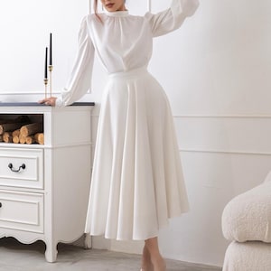 Op de afbeelding: Een witte tweedelige outfit met een blouse met lange mouwen en een hoge halslijn en een vloeiende midi-rok. De blouse heeft pofmouwen. De outfit wordt gedragen met doorzichtige hakken. Een witte dressoir en zwarte kaarsen staan op de achtergrond.